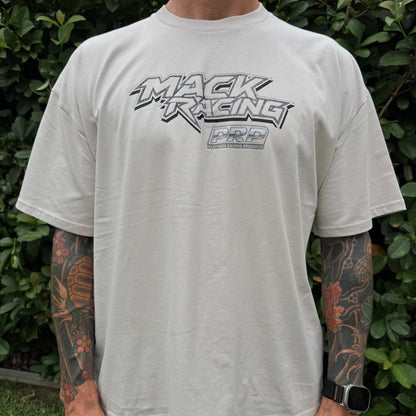 Person wearing a white t-shirt with 'Mack Racing' logo in front of green foliage
